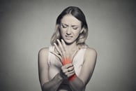Young woman holding her painful wrist isolated on gray wall background. Sprain pain location indicated by red spot. Negative face expression-1